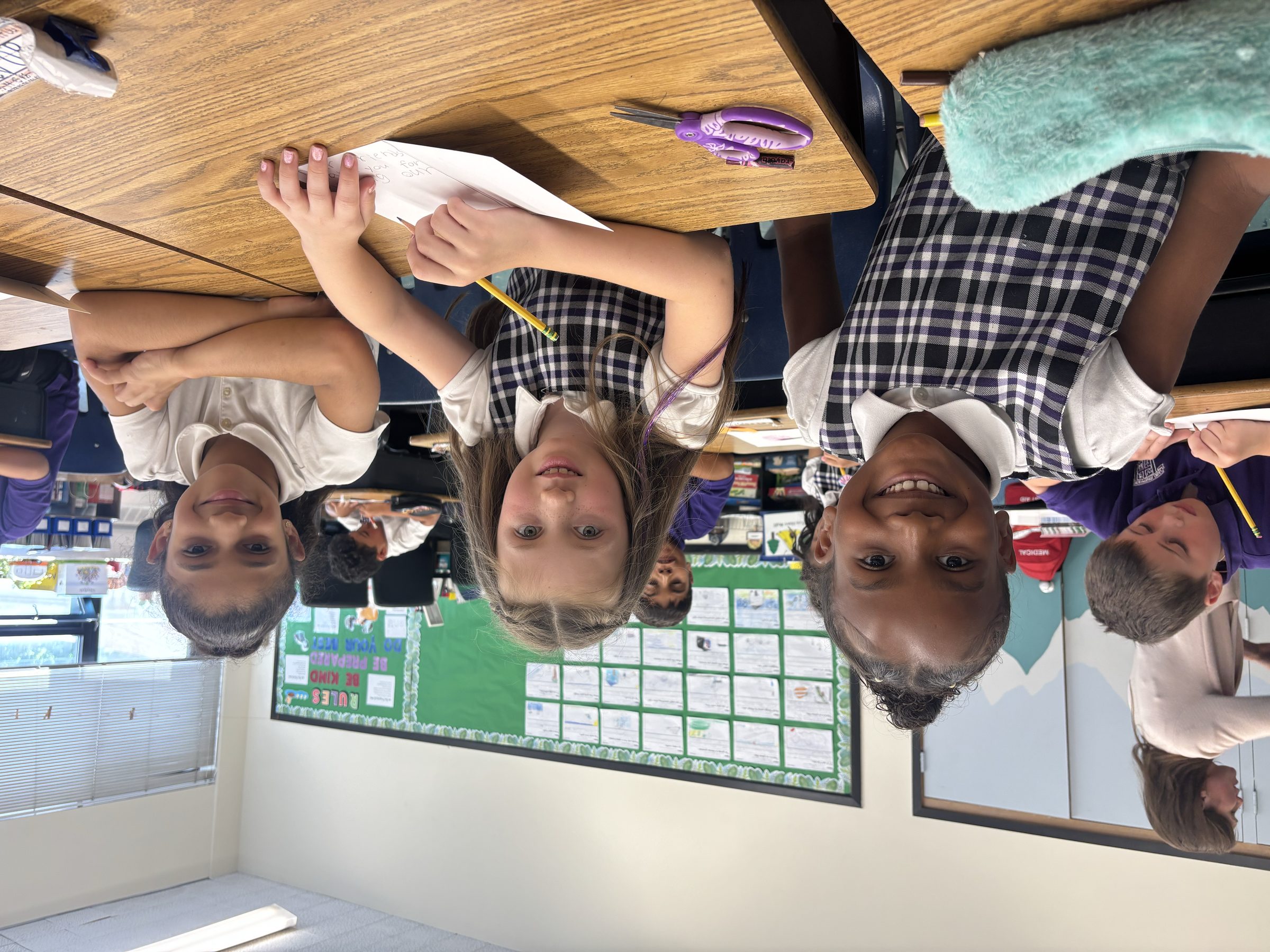Three students in uniform smiling at their desks in a Catholic school classroom, one of them writing with a pencil